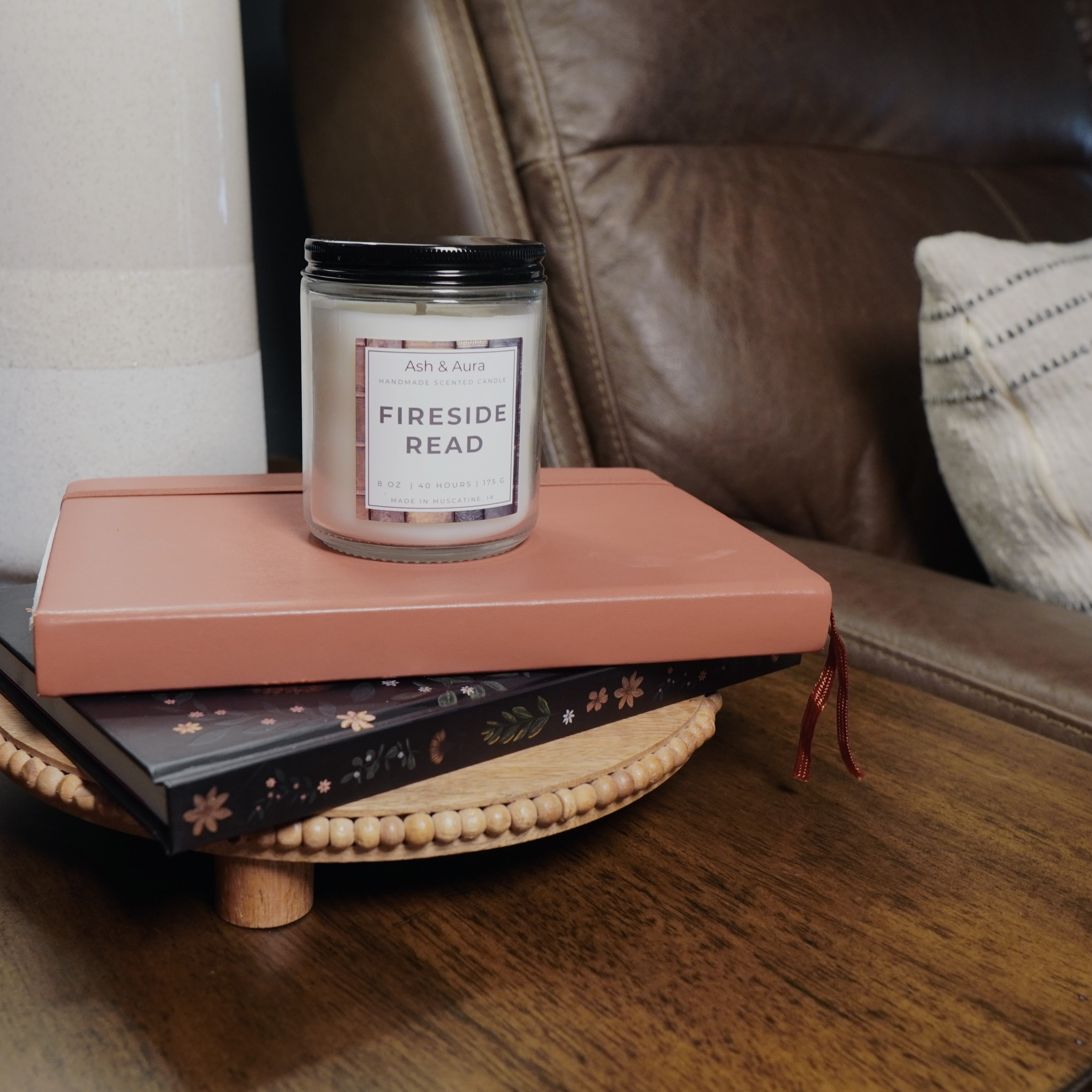 A lit Fireside Read candle sitting on a wooden table next to a stack of books and a pair of glasses.